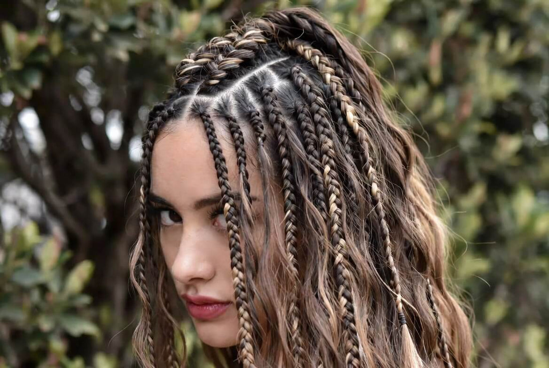 A striking close-up of a woman with a mix of small braids and loose, wavy hair in dark brown with blonde highlights. The braids are artfully arranged from a center part, with some strands framing her face.