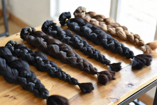 Several long, pre-braided hair extensions in shades of black, dark brown, and light brown laid out on a wooden table. Some braids have gold rings at the top, ready for installation.