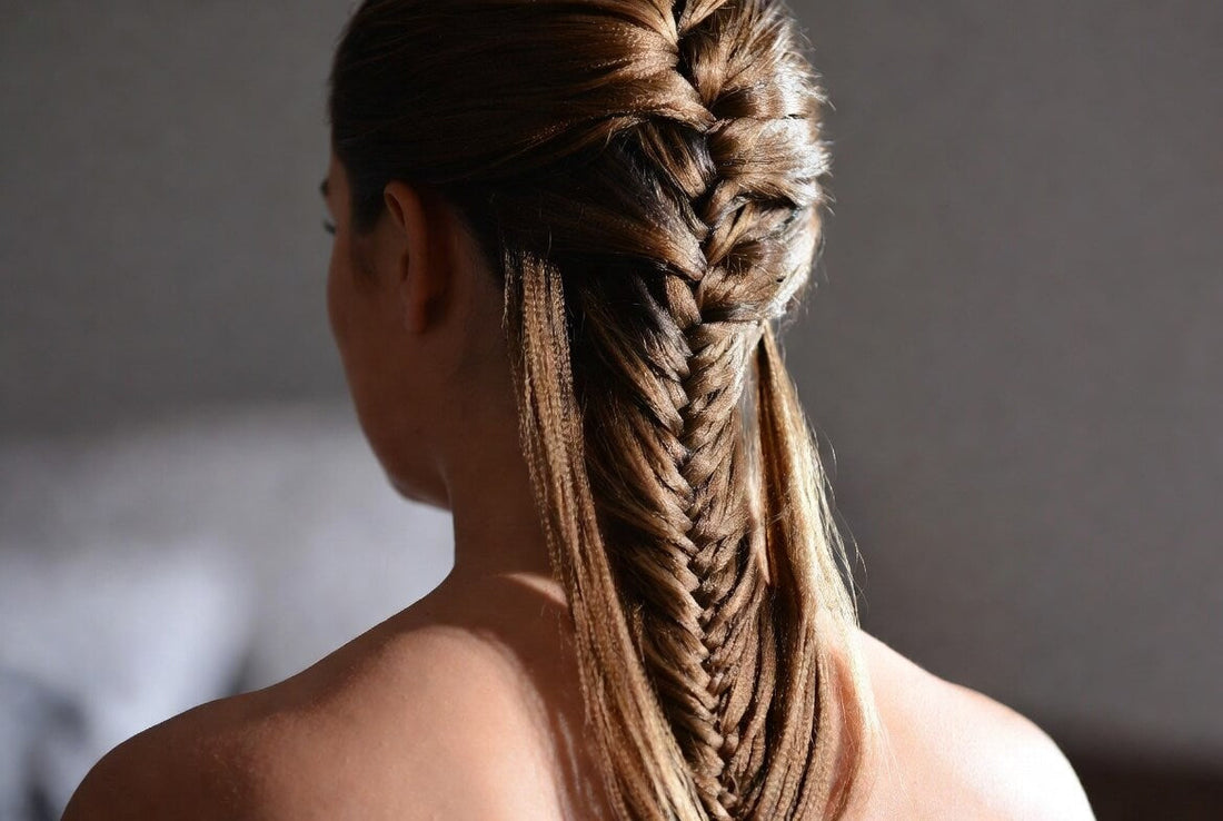 A woman viewed from the back, showcasing an intricate, thick Dutch braid that starts at the crown and transitions into a low ponytail with loose strands. Her smooth brown hair is styled in a voluminous, textured braid against a soft indoor background.