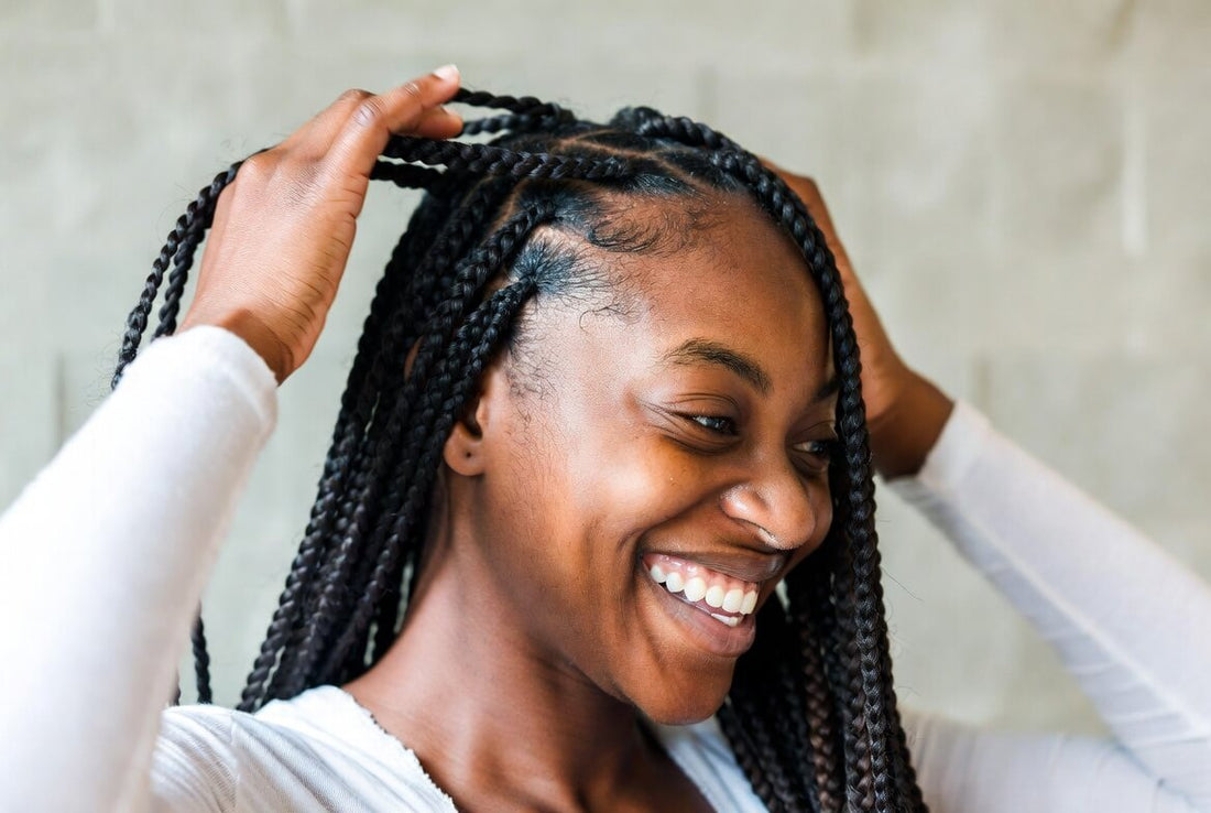 A smiling young Black woman with long, neat box braids and a criss-cross parting on top. She gently touches her hair with one hand, looking down with a soft, confident expression.