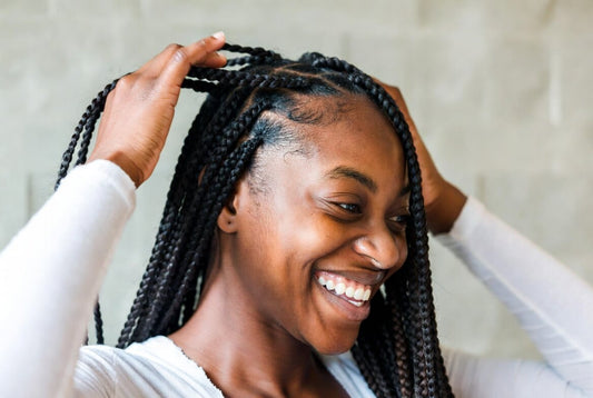 A smiling young Black woman with long, neat box braids and a criss-cross parting on top. She gently touches her hair with one hand, looking down with a soft, confident expression.