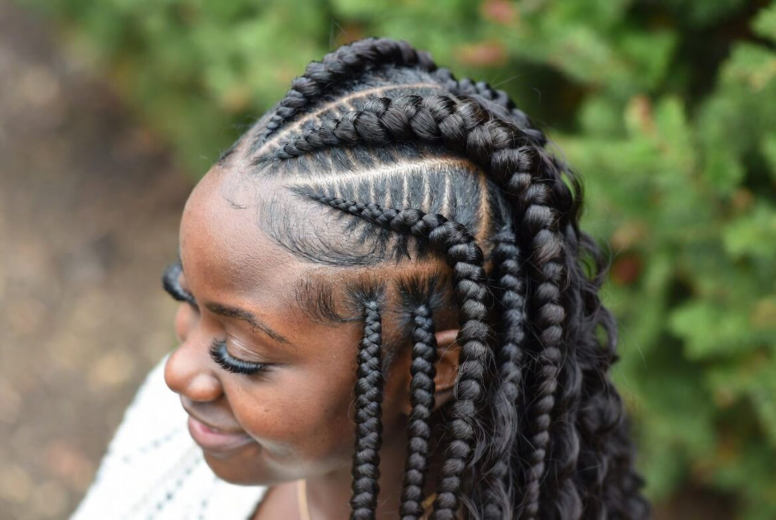 Close-up side profile of a woman with neatly parted cornrow braids that transition into longer individual braids. The intricate pattern is visible on her scalp, and she has long eyelashes and a slight smile.