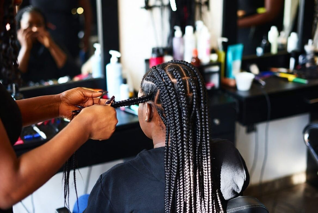A professional hairstylist’s hands actively braiding a client’s hair in a salon setting. The client is seated with her back to the camera, showing partially completed tight cornrows and box braids. Salon mirrors and products are visible in the background.