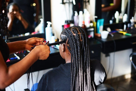 A professional hairstylist’s hands actively braiding a client’s hair in a salon setting. The client is seated with her back to the camera, showing partially completed tight cornrows and box braids. Salon mirrors and products are visible in the background.