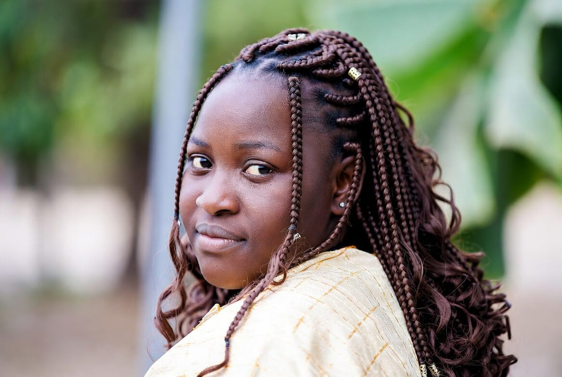Close-up portrait of a young woman with long medium knotless box braids in dark brown and blonde highlights, adorned with small gold hair charms. She is smiling softly while looking over her shoulder in a bright indoor setting.
