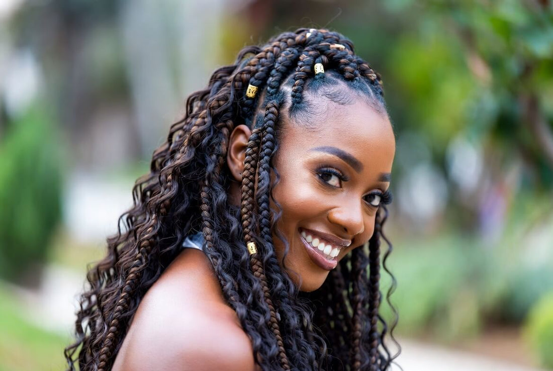 Radiant portrait of a smiling woman with long goddess braids — a mix of cornrows and loose voluminous curls with gold beads. She has a joyful expression and is looking slightly to the side outdoors.