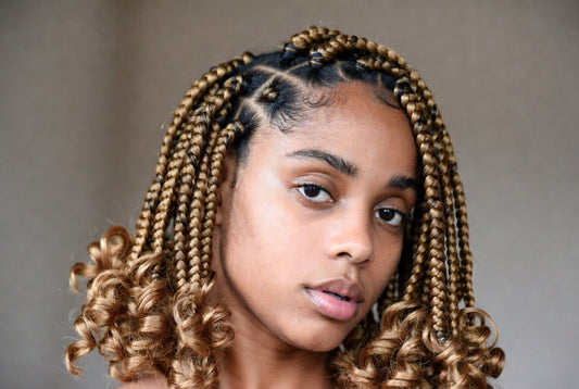 Close-up of a young woman with honey-blonde knotless braids that transition into soft, defined curly ends. She has a neutral expression and is looking directly at the viewer.