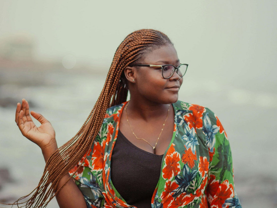 Woman with long reddish-brown lemonade braids wearing glasses and a colorful floral top, posing by the water.