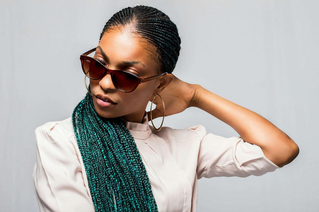 Confident woman with teal-colored braids wearing sunglasses and gold hoops, posing with hand behind her head.